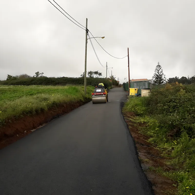 Camino rural recién asfaltado entre fincas con un rodillo al fondo