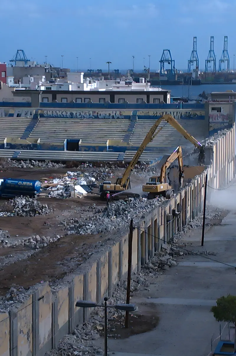 Excavadora derribando un graderío con las grúas del puerto al fondo